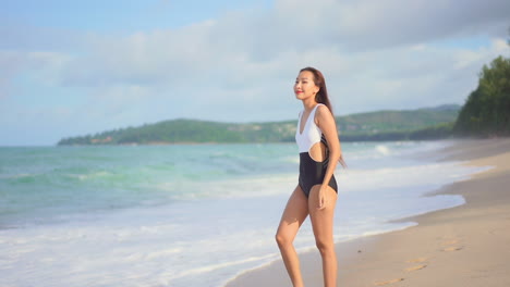 Young-Asian-woman-wearing-black-and-white-swimming-suit-walking-along-the-beach-line-of-the-picturesque-island,-following-shot,-slow-motion