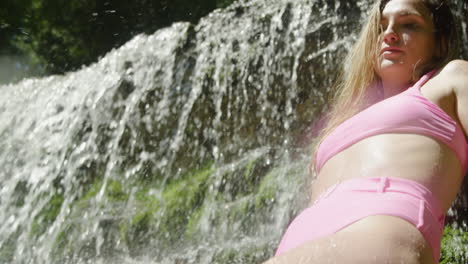 Junge-Frau-Im-Bikini-Posiert,-Während-Sie-Auf-Den-Felsen-Unter-Einem-Kleinen-Wasserfall-Sitzt