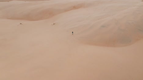 High-aerial-view-of-sandboarder-walking-alone-on-big-sand-dunes-near-the-tropical-beach-of-Garopaba,-Santa-Catarina,-Brazil