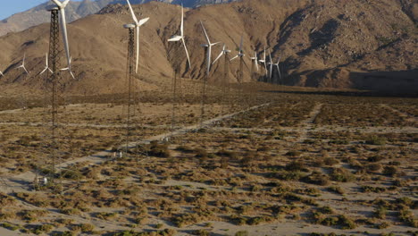 Stunning-aerial-view-of-drone-flying-backwards-alongside-many-wind-turbines-with-huge-mountain-in-the-background-at-wind-farm-near-Palm-Springs-in-the-Mojave-Desert,-California,-USA