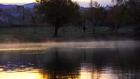 Water-lake-steaming-at-morning-on-city-park,-people-walking-around-trees