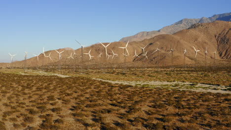 Drone-flying-towards-dozens-of-wind-turbines-at-a-huge-wind-farm-with-mountains-in-the-background-at-near-Palm-Springs-in-the-Mojave-Desert,-California,-USA