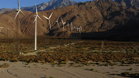 Aerial-view-panning-from-left-to-right-overlooking-aligned-wind-turbines,-desert-and-huge-mountains-near-Palm-Springs-in-the-Mojave-Desert,-California,-USA