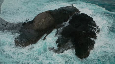Waves-crashing-on-isolated-stretch-of-rocks-surrounded-by-water,-aerial