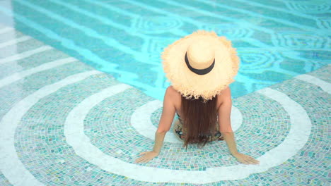 Backside-view-of-pretty-woman-in-the-tropical-spa,-sitting-at-the-pool,-wearing-hat-and-swimsuit,-top-view