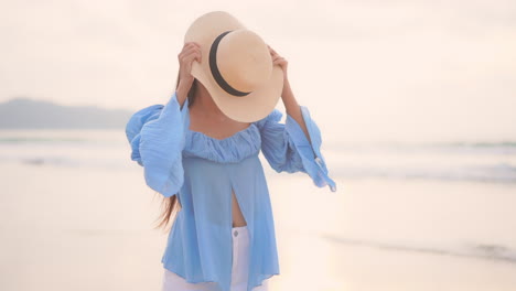 A-pretty-young-woman-puts-her-sunhat-on-as-she-walks-along-the-edge-of-a-sandy-shoreline