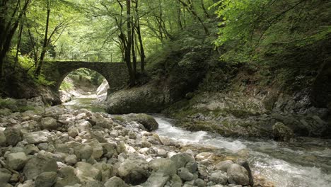river-flowing-in-secluded-idyllic-canyon,-stone-bridge-crossing,-lush-greenery