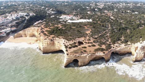 Aerial,-pull-back-reveal,-drone-footage-of-the-unique-cliffs-and-rock-formations-along-the-shoreline-near-the-coastal-village-of-Benagil,-Portugal