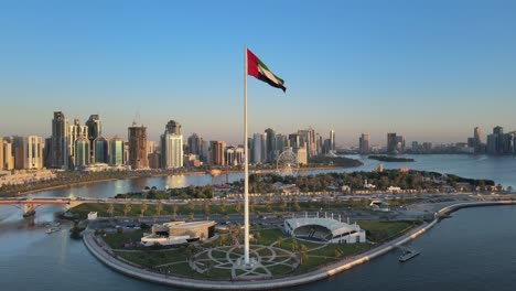 Drohnenansicht-Der-Flagge-Der-Vereinigten-Arabischen-Emirate,-Die-In-Der-Luft-Weht,-Der-Blaue-Himmel-Im-Hintergrund,-Das-Nationale-Symbol-Der-Vereinigten-Arabischen-Emirate-über-Der-Flaggeninsel-Von-Sharjah,-Vereinigte-Arabische-Emirate,-Nationalfeiertag-Der-Vereinigten-Arabischen-Emirate,-4k