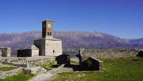 Historic-clock-tower-of-Gjirokastra's-castle-and-ruined-stone-walls-with-mountain-background