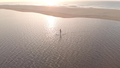 Aerial-shot-orbiting-around-a-man-on-a-stand-up-paddle-board-on-a-lake-near-the-ocean-with-the-sun-rising-above-the-horizon-on-the-coast-in-southern-Australia