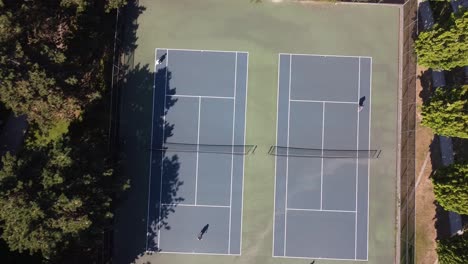 aerial-birds-eye-view-over-tennis-courts-surrounded-by-a-park-by-a-local-hometown-road-with-empty-steps-by-a-dry-futuristic-fountain-water-sprinklers-in-the-summer-where-its-sunny-lush-green-grass-1-3