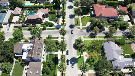 Drone-follows-white-and-silver-cars,-separately-and-together,-driving-up-beautiful-palm-tree-and-mansion-lined-street-in-classic-West-Hollywood,-Los-Angeles,-Callifornia-neighborhood