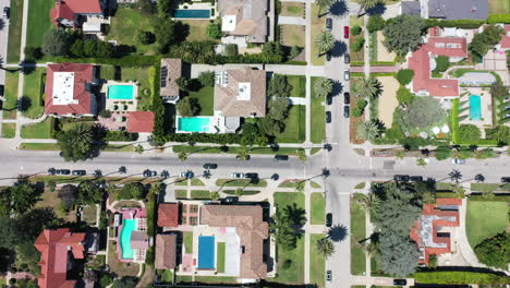 Drone-shot-flying-over-beautiful-palm-tree-lined-street-following-black-car-driving,-the-street-is-surrounded-by-mansions-with-swimming-pools-in-West-Hollywood,-Los-Angeles,-California