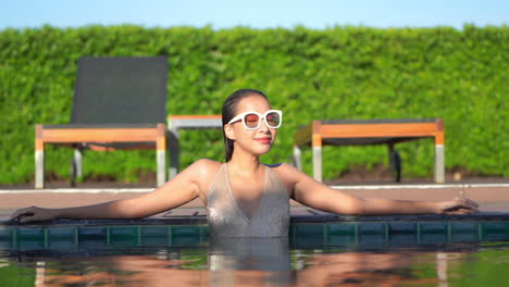 Attractive-woman-in-shimmering-gold-swimsuit-and-large-sunglasses-standing-in-swimming-pool-turns-head-and-looks-toward-camera-with-seductive-smile