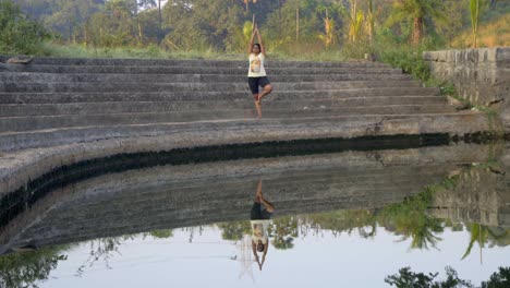 girl-doing-tree-pose-vrhsasana-double-image-reflection-in-still-water-on-steps-of-lade
