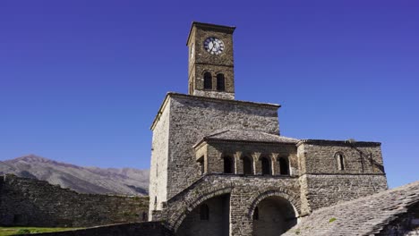 Cinematic-shot-of-clock-tower-with-stone-and-arched-walls-of-Argjiro-Castle-in-blue-bright-sky-background