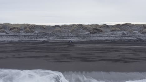 Lone-quad-biker-travels-on-scenic-black-beach-in-cold-Iceland,-truck-right