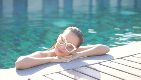 Gesicht-Nahaufnahme-Einer-Schönen-Asiatischen-Frau-In-Weißer-Sonnenbrille,-Die-Sich-Tagsüber-Im-Schwimmbad-Entspannt-Und-Ihren-Kopf-Auf-Gesperrte-Hände-Legt