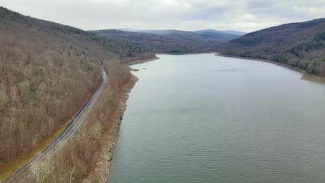 Drone-flying-forward-over-a-mountain-lake-alongside-a-mountain-road-in-early-winter,-over-a-mountain-valley