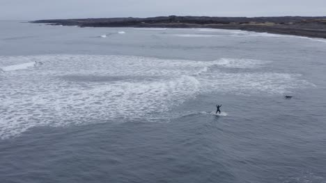 Surfer-balancing-on-surfboard-while-riding-small-wave-at-Sandvík-beach,-aerial