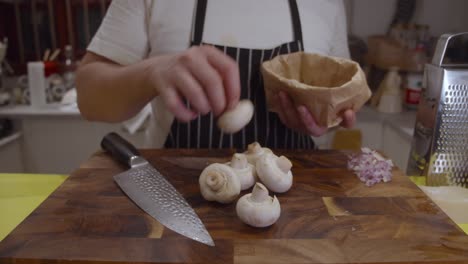 Chef-puts-out-white-mushrooms-onto-wooden-cut-board-from-paper-bag-in-the-kitchen