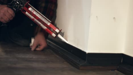 Grout-sealent-out-of-silicon-with-a-white-color-being-applied-on-a-skirting-base-board-in-a-new-apartment-by-a-crafts-person-with-a-cartridge-gun-on-a-black-wooden-floor