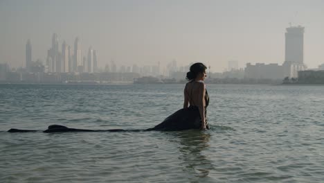 Wunderschöne-Frau-In-Schwarzem-Kleid-Geht-Ins-Meer-Mit-Der-Skyline-Von-Dubai-Im-Hintergrund---Statisch,-Weitwinkelaufnahme