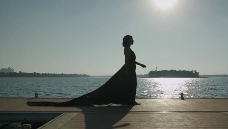 Beautiful-Woman-In-Long-Black-Dress-Walks-Across-Wooden-Deck-Near-The-Sea---Static,-Wide-Shot