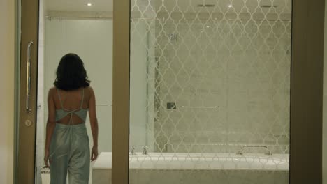 Sexy-Woman-Removes-Knot-From-Back-Of-Jumpsuit-And-Walks-Into-Bathroom-Then-Sits-Near-The-Tub---Static