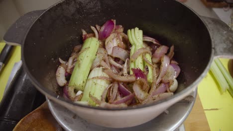 Stirring-onion-slices-mixed-with-celery-and-spices-with-wooden-kitchen-spoon-inside-deep-cooking-pot
