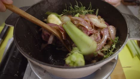 Stirring-beef-shank-mixed-with-onion-slices,-celery,-spices-with-wooden-spoon-inside-deep-cooking-pot