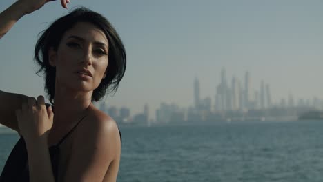 Schöne-Frau-Im-Schwarzen-Kleid-Posiert-Im-Freien-Mit-Nebliger-Skyline-Von-Dubai-Marina-Im-Hintergrund---Mittlere-Nahaufnahme