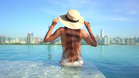 A-young-woman-with-her-back-to-the-camera-sits-on-the-edge-of-a-rooftop-resort-infinity-pool,-adjusts-her-sun-hat-while-taking-in-the-urban-skyline-in-the-distance