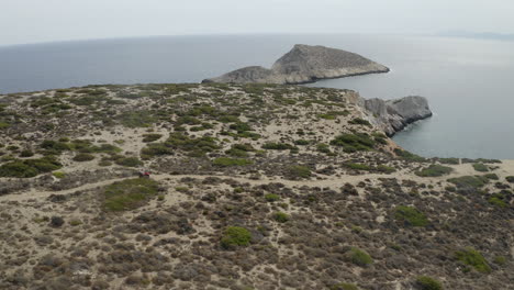 Couple-on-quad-bike,-ATV-off-roading-on-rough-rocky-road-track-near-sea-on-Ios,-Greece-drone-aerial-shot