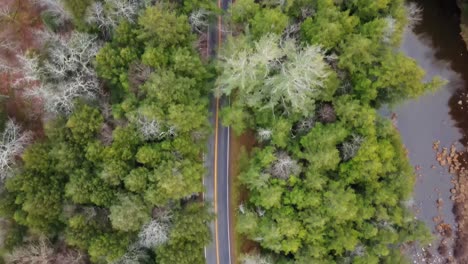 Birds-eye-drone-video-of-a-mountain-road,-bare-deciduous-trees,-coniferous-pine-trees,-and-a-small-river-in-early-winter-in-the-mountains