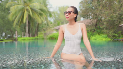 An-attractive-young-woman-in-a-one-piece-bathing-suit-sits-along-the-shallow-edge-of-a-resort-pool