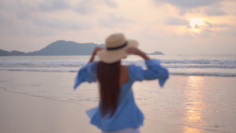 Young-woman-walks-on-seashore-at-sunset-and-raises-her-arms