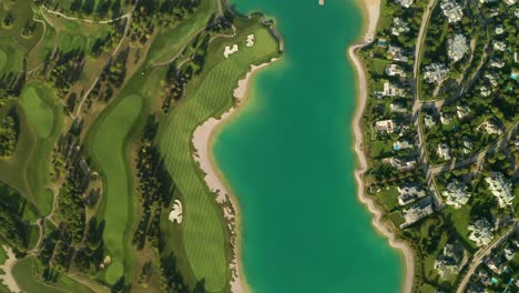 Aerial-overhead-view-of-a-turquoise-lake-next-to-a-golf-resort-with-beautiful-abstract-patterns,-sand-traps,-trees-tennis-courts,-and-a