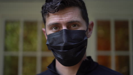 Young-man-puts-on-a-black-face-mask-to-protect-himself-and-others-from-a-virus-then-looks-at-the-camera---2020-global-pandemic