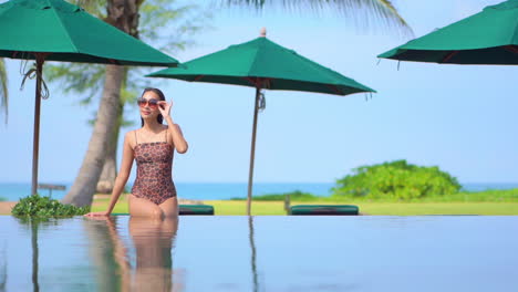 Vacation-on-Exotic-Tropical-Destination,-Pretty-Asian-Female-Sitting-on-Infinity-Pool-Edge-in-Swimsuit-With-Green-Foliage-and-Sea-in-Background,-Full-Frame