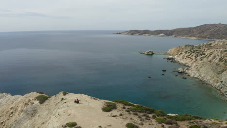 Revealing-drone-shot-of-beautiful-scenic-coastal-cliffs-in-Ios,-Greece-with-Couple-on-quad-bike