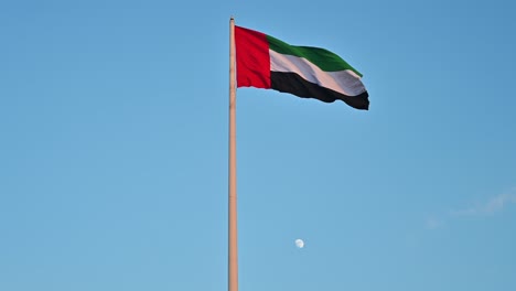 Die-Flagge-Der-Vae-Weht-In-Der-Luft,-Der-Mond-Des-Ersten-Viertels-Im-Hintergrund,-Das-Nationale-Symbol-Der-Vereinigten-Arabischen-Emirate,-Der-Nationaltag-Der-Vae-2020,-4k-video