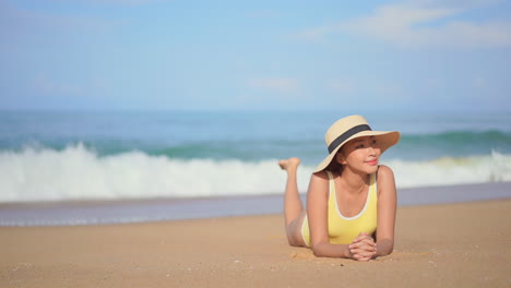 Asian-girl-wearing-yellow-swimsuit-and-big-hat-lying-on-beach-smiling-looking-at-camera