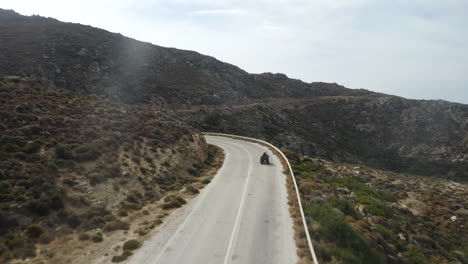 Fast-flying-drone-following-couple-on-ATV,-quad-bike-in-the-mountains-of-Ios,-Greece