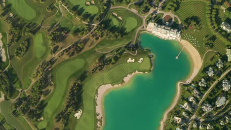 Aerial-overhead-view-of-a-turquoise-lake-next-to-a-golf-resort-with-beautiful-abstract-patterns,-sand-traps,-trees-tennis-courts,-and-a