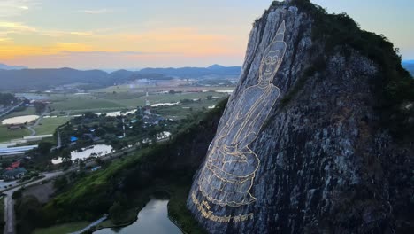 4k-drone-footage-circling-an-image-of-Buddha,-sitting-cross-legged,-engraved-with-gold-into-the-northern-face-of-a-limestone-hill-in-Khao-Chi-Chan,-Thailand