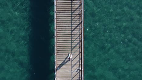 Rising-aerial-shot,-looking-down-on-a-little-girl-walking-along-a-wood-pier-by-the-sea