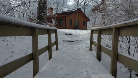 Walking-Over-Bridge-Towards-Cabin-in-Winter-Snowy-Landscape,-Forward-Dolly