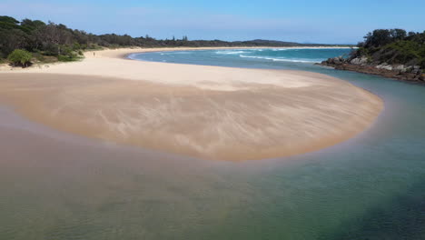 Wide-rotating-drone-shot-of-the-South-Pacific-Ocean-and-Korogoro-Creek-with-wind-blowing-sand-across-a-sand-bar-at-Hat-Head-New-South-Wales,-Australia
